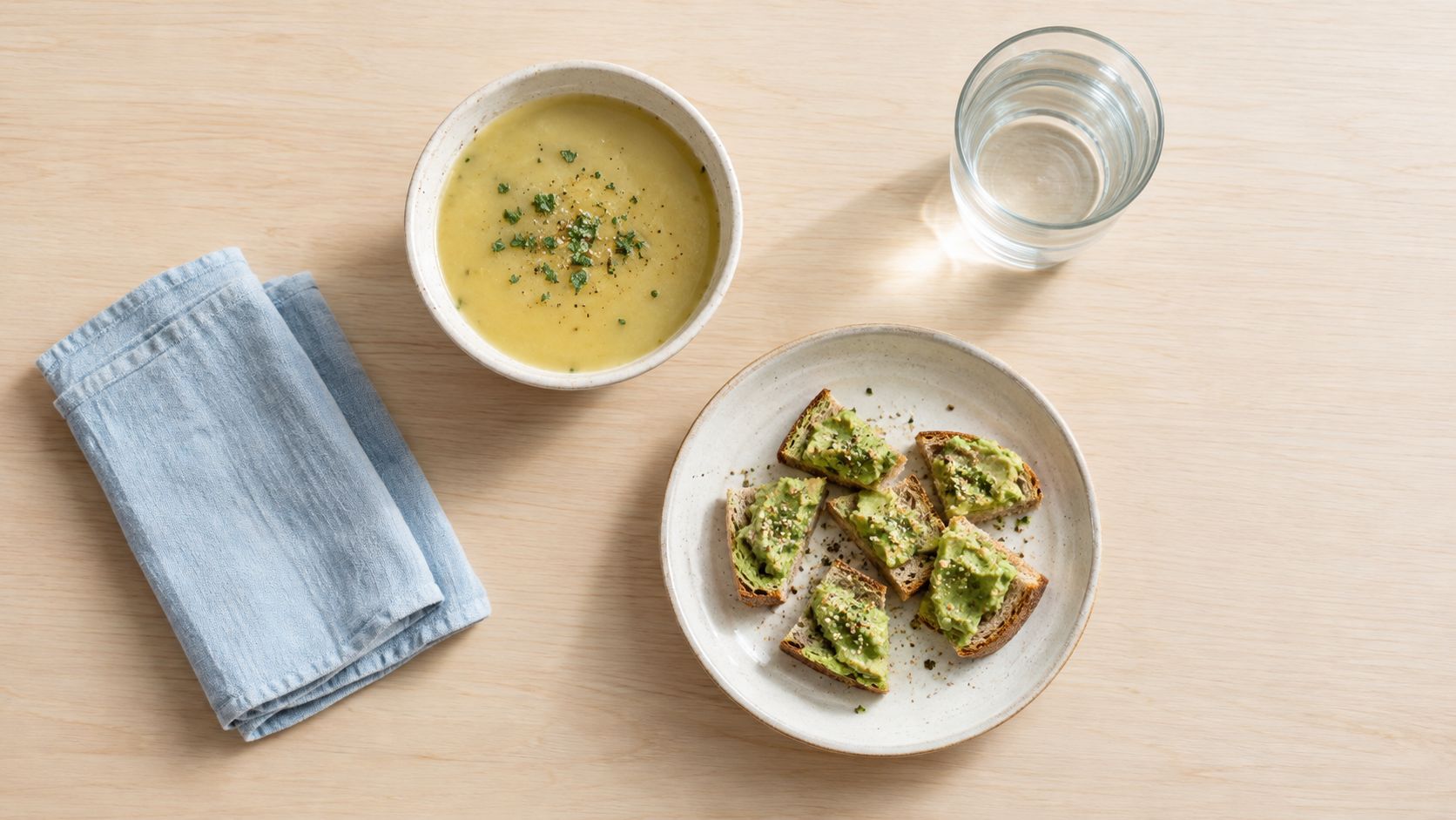 A light meal of potato soup with herbs and small avocado toast slices on a wooden table.