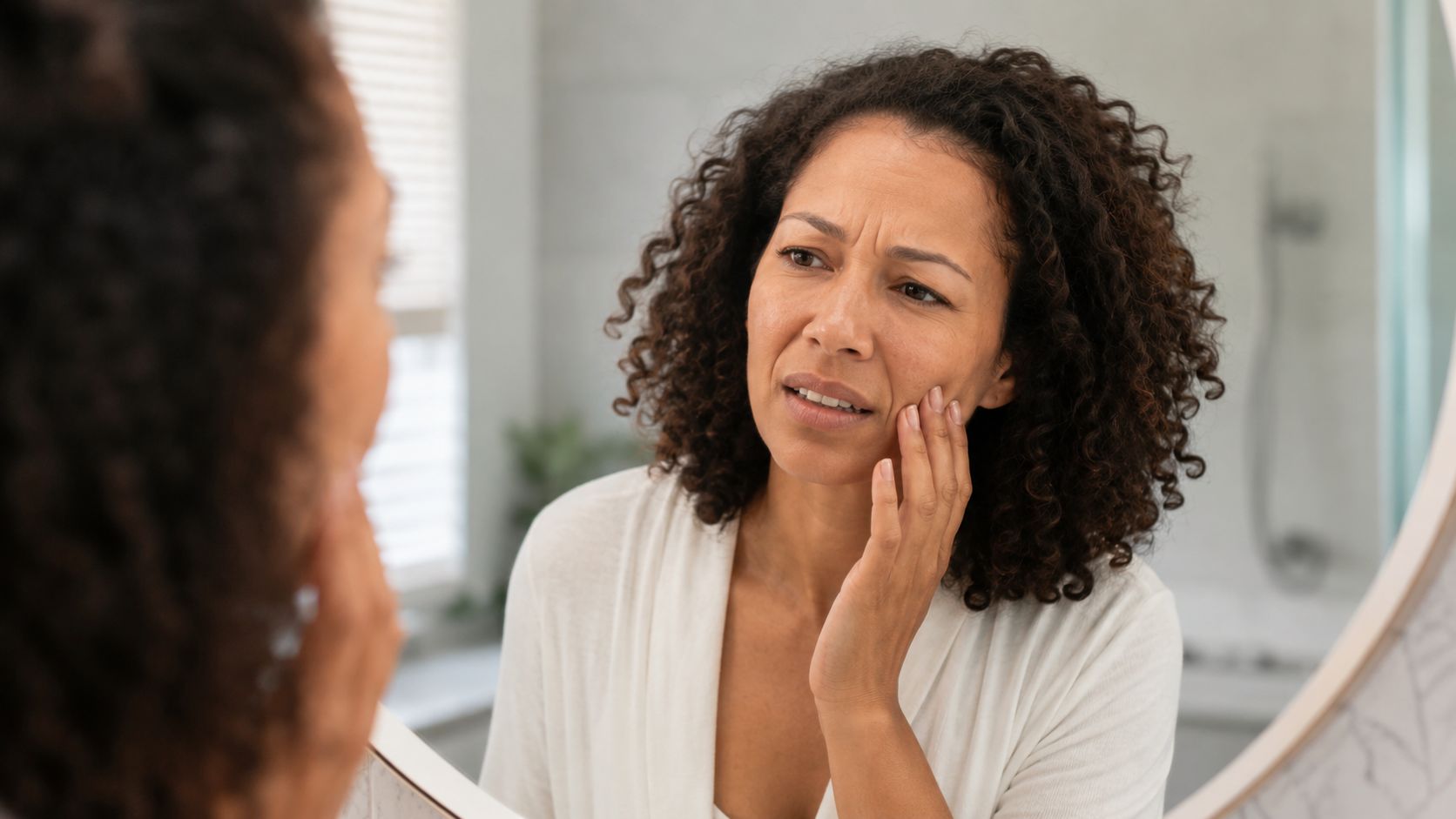 A concerned woman checking her teeth and facial skin in a bathroom mirror reflection.