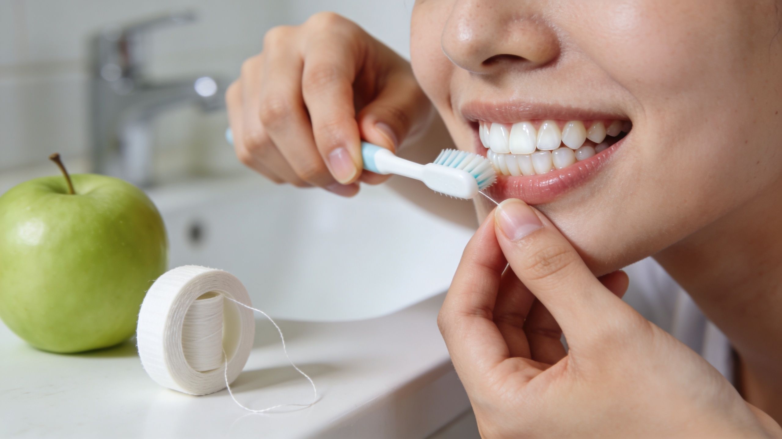 A young woman brushing her teeth and using dental floss in a bright, modern bathroom setting.