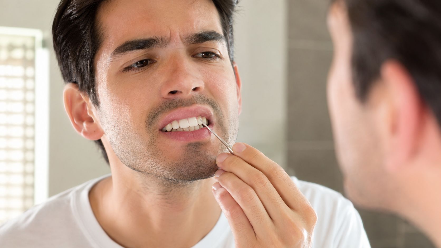 A man examines his teeth in a bathroom mirror using a small metal dental tool.