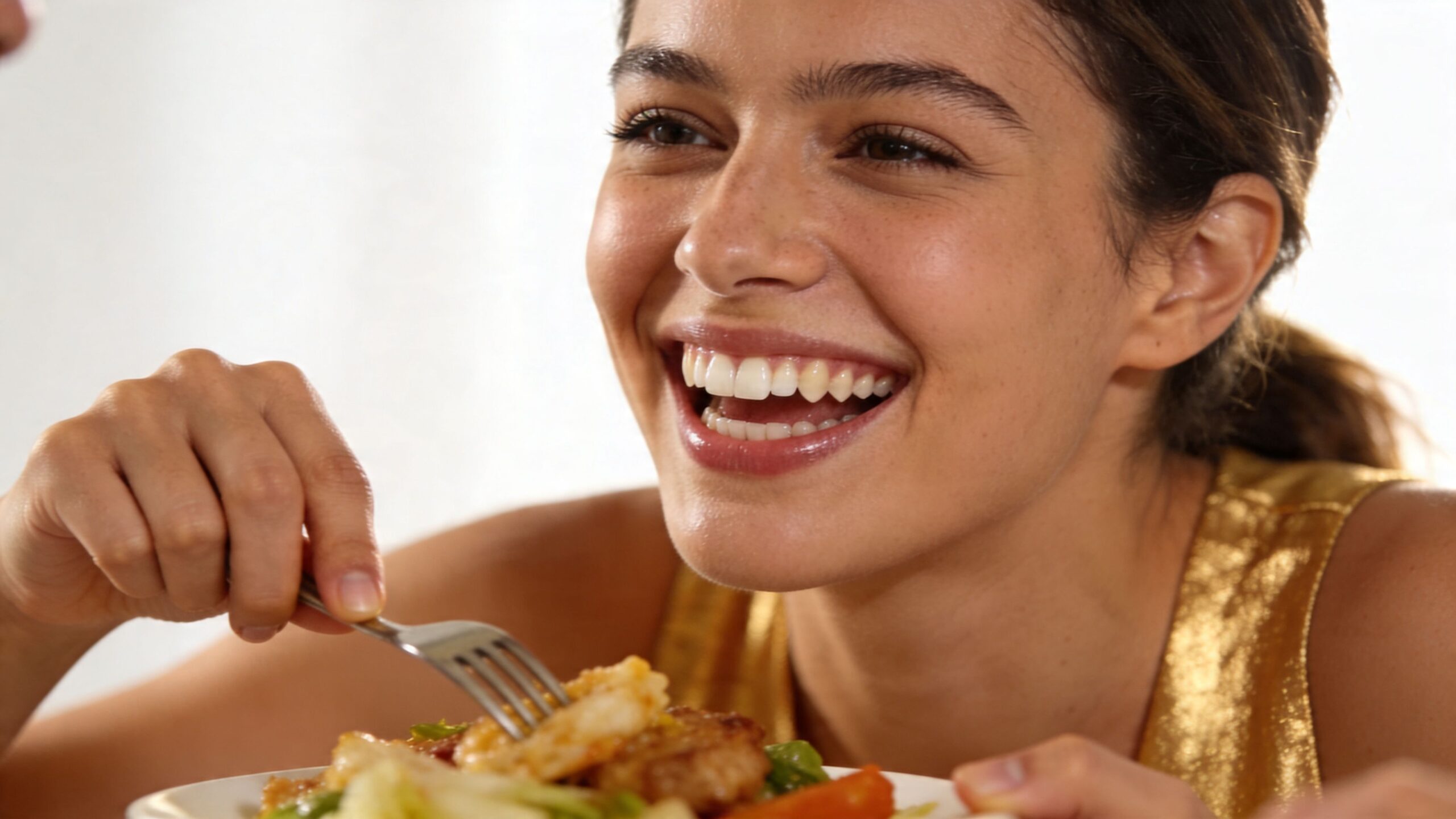 A happy young woman smiling while eating a delicious meal with a fork from a white plate.
